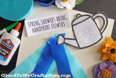 A craft scene with a glue bottle, blue and yellow felt flowers, blue crepe paper strips, a paper watering can, and a card reading “Spring Showers Bring Handprint Flowers,” features fun Spring Printables on a white background.