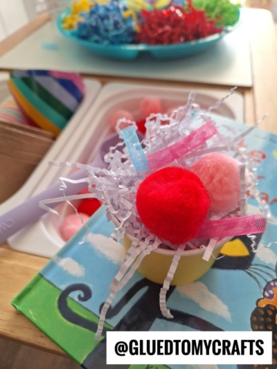 A small yellow bowl filled with colorful pom-poms, paper shreds, and ribbons sits on a childrens craft book. Craft supplies and shredded paper are visible in the background. Text overlay reads @GLUEDTOMYCRAFTS.