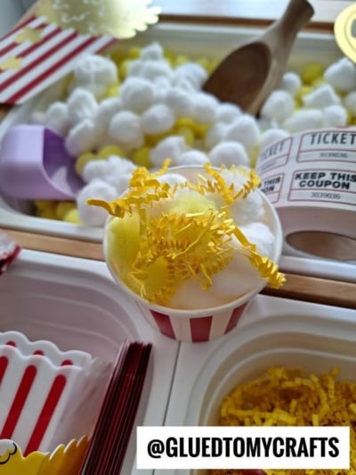 A small striped cup filled with cotton balls and yellow paper shreds sits on a tray, surrounded by pretend popcorn, tickets, and scoops for a craft activity. @GLUEDTOMYCRAFTS is written at the bottom.