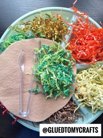 A round tray holds colorful shredded paper, a brown felt circle, and plastic tweezers, arranged for a craft activity. Text at the bottom reads @GLUEDTOMYCRAFTS.