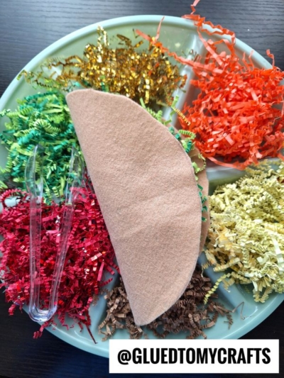 A felt taco shell sits on a plate surrounded by colorful shredded paper representing taco fillings; a clear plastic fork and a label reading @GLUEDTOMYCRAFTS are also on the plate.