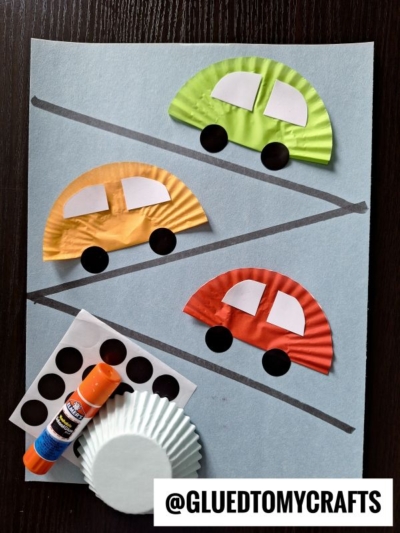 Three paper cars made from colored cupcake liners, paper circles, and rectangles are arranged on a blue sheet with black diagonal lines like a road. Glue, black dot stickers, spare cupcake liners, and coding label crafts sit nearby.