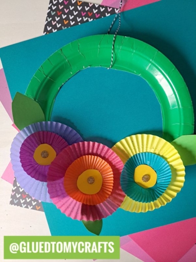 A colorful cupcake liner flower wreath made from a paper plate, decorated with three paper cupcake liner flowers and green leaves, displayed on vibrant sheets of paper. The handle features black-and-white twine. @GLUEDTOMYCRAFTS is at the bottom.
