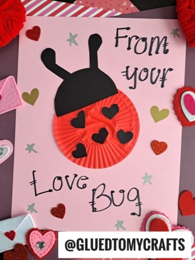 A handmade Valentine’s card features a ladybug made from a red cupcake liner and black paper on pink paper, decorated with hearts and the text “From your Love Bug.” This adorable cupcake liner card craft is surrounded by various felt hearts.