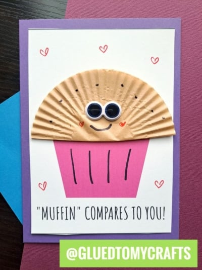 A handmade cupcake liner card features a cupcake with a brown muffin liner top, googly eyes, a smile, and a pink paper base. Red hearts and the text Muffin compares to you! add charm to this cute card craft.