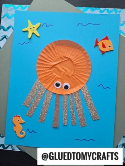 A children’s jellyfish craft made with an orange cupcake liner for the body—perfect for cupcake liner card crafts—googly eyes, glittery paper tentacles, and foam sea creature stickers on a blue background. @GLUEDTOMYCRAFTS is written below.