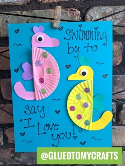 Two seahorses made from painted paper plates, googly eyes, and paper shapes are glued to blue paper with handwritten text reading Swimming by to say I love you! and hearts—perfect inspiration for cupcake liner card crafts. @GLUEDTOMYCRAFTS is at the bottom.