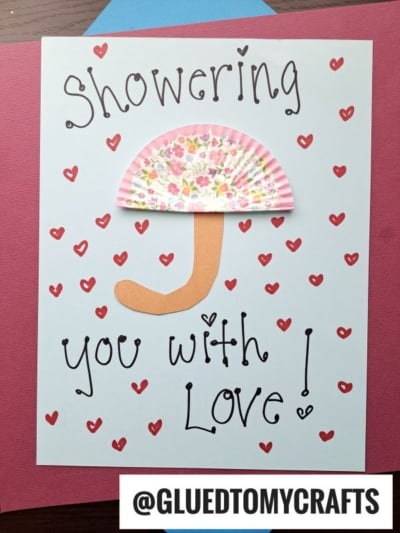 Handmade cupcake liner card crafts with red hearts, a cupcake liner shaped like an umbrella, and "Showering you with love!" in playful font. The handle is a paper cutout. @GLUEDTOMYCRAFTS appears at the bottom.