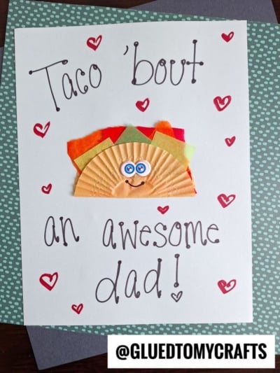 Handmade cupcake liner card crafts "Taco bout an awesome dad!" in playful letters, decorated with red hearts. A smiling taco made from a cupcake liner and colored paper is in the center. @GLUEDTOMYCRAFTS is at the bottom.