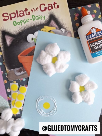 A children’s book titled Splat the Cat: Oopsie-Daisy sits next to a blue paper with daisy crafts made from cotton balls and yellow paper, a bottle of Elmer’s glue, and polka dot stickers. @GLUEDTOMYCRAFTS is displayed at the bottom.