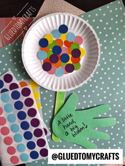 A handmade craft with colorful dot stickers on a paper plate, green paper cut as a hand and stem, plus a coding label crafts touch. The message “A little hand, a big bloom!” appears on patterned star paper. @GLUEDTOMYCRAFTS is at the bottom.