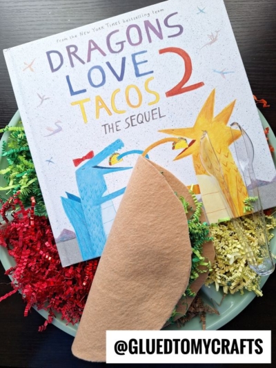 A children’s book titled Dragons Love Tacos 2: The Sequel sits on a plate with red and yellow crinkle paper, a plastic fork, and a felt taco shell. @GLUETOMYCRAFTS is written at the bottom.