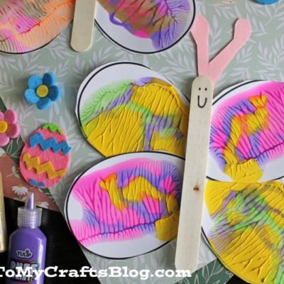 Colorful butterfly craft made with oval, squished paint wings, a popsicle stick body with a smiley face, and pink paper antennae. Felt flowers, Easter eggs, and squeeze bottles of puffy paint are nearby.