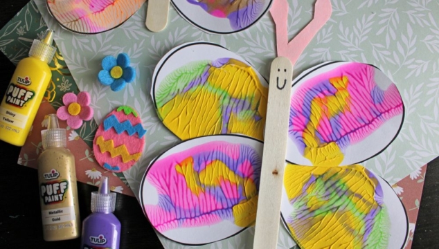 Colorful butterfly craft made with oval, squished paint wings, a popsicle stick body with a smiley face, and pink paper antennae. Felt flowers, Easter eggs, and squeeze bottles of puffy paint are nearby.