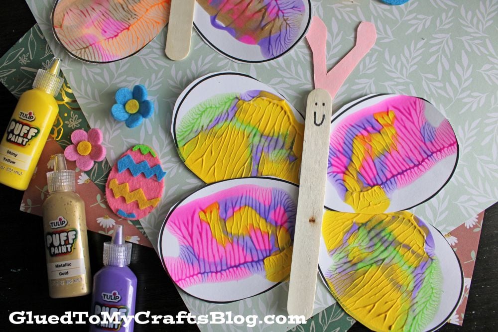Colorful butterfly craft made with oval, squished paint wings, a popsicle stick body with a smiley face, and pink paper antennae. Felt flowers, Easter eggs, and squeeze bottles of puffy paint are nearby.