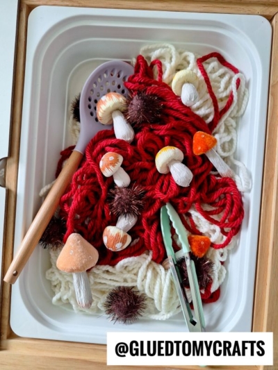 A sensory bin filled with red and white yarn, fake mushrooms, brown spiky balls, a purple spoon, and green tongs. The bottom has a label reading @GLUEDTOMYCRAFTS.