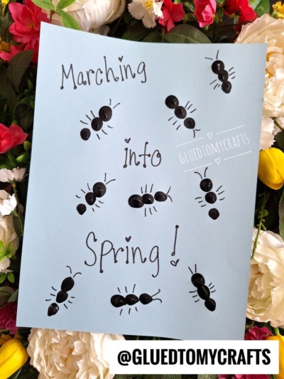 A blue paper with Marching into Spring! written in black, surrounded by ant drawings made from thumbprints and pen—a fun idea for THUMBPRINT CRAFTS FOR KIDS. The paper rests on colorful flowers, with @GLUEDTOMYCRAFTS printed at the bottom.
