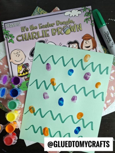 A pastel green paper with green zigzag lines and colorful fingerprint dots sits on a table with a Charlie Brown Easter book, paint pots, a paintbrush, a Sharpie marker, and @GLUEDTOMYCRAFTS label—perfect for thumbprint crafts for kids.
