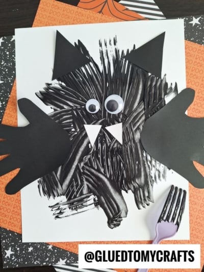 A Halloween cat craft made with black paint, fork strokes, googly eyes, black paper ears and paws, and white paper fangs, displayed on layered Halloween-themed paper. A purple plastic fork is beside the artwork.