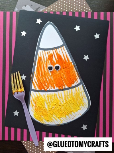 A candy corn craft made with orange and yellow paint textured using a fork, two googly eyes, and a background of black paper with white stars. A purple fork lies next to the craft. The text reads @GLUEDTOMYCRAFTS.
