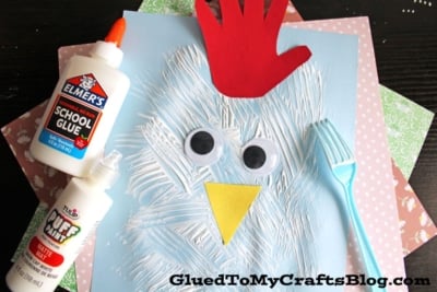 A children's chicken craft made from a blue paper, white painted strokes, two large googly eyes, a yellow paper beak, a red handprint for a comb, and a blue plastic fork, displayed on colorful patterned sheets.