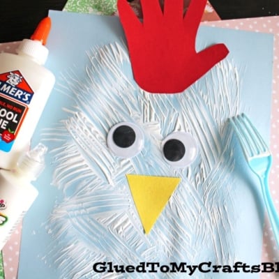 A children's chicken craft made from a blue paper, white painted strokes, two large googly eyes, a yellow paper beak, a red handprint for a comb, and a blue plastic fork, displayed on colorful patterned sheets.