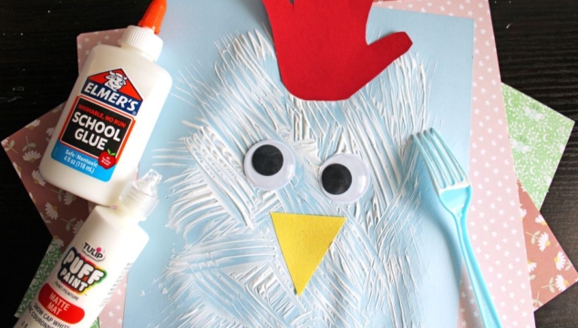 A children's chicken craft made from a blue paper, white painted strokes, two large googly eyes, a yellow paper beak, a red handprint for a comb, and a blue plastic fork, displayed on colorful patterned sheets.