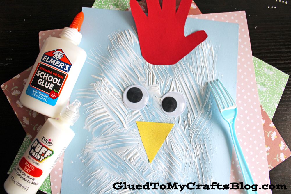 A children's chicken craft made from a blue paper, white painted strokes, two large googly eyes, a yellow paper beak, a red handprint for a comb, and a blue plastic fork, displayed on colorful patterned sheets.
