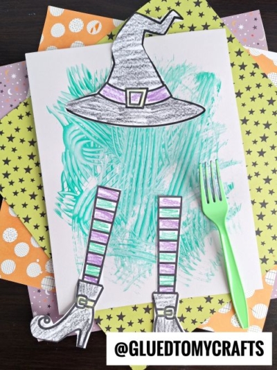 A witch-themed craft shows a paper witch hat, legs, and shoes, colored with purple and gray, placed on a scribbled green background. A green plastic fork and star-patterned papers are in the background.