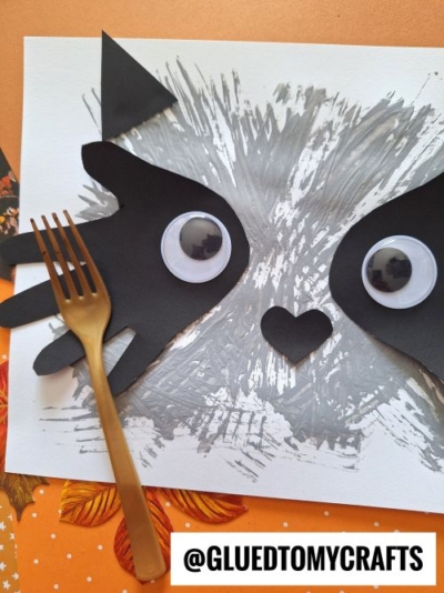 A cat face is crafted from gray paint handprints, black paper cutouts for ears and paws, large googly eyes, and a heart-shaped nose. A gold plastic fork and autumn leaves are nearby. The image has the text @GLUEDTOMYCRAFTS.