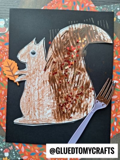 A paper squirrel craft on black paper, decorated with brown coloring, gold star-shaped glitter, a googly eye, and an orange paper leaf. A purple plastic fork is used to create texture on the tail.