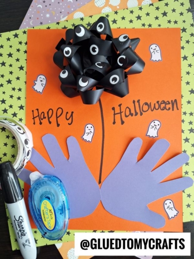 A Halloween craft features an orange paper with Happy Halloween, two purple handprints as flower stems, a black bow with eyes as the flower, ghost stickers, a Sharpie, tape runner, and star-patterned paper.