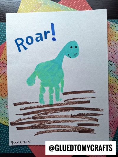 Children’s HANDPRINT FATHER'S DAY painting of a green dinosaur with “Roar!” above. The dinosaur’s body and head are made from a blue handprint, standing on brown lines for ground. ©GluedToMyCrafts appears at the bottom.
