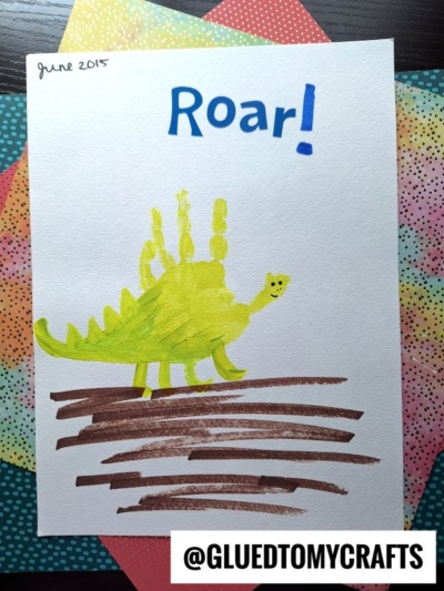 A child’s dinosaur painting features a yellow-green stegosaurus made from a handprint, perfect for HANDPRINT FATHER'S DAY crafts. "Roar!" is written in blue at the top, on white paper with colorful patterned paper beneath.