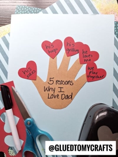 A HANDPRINT FATHER'S DAY paper craft with red heart cutouts on each finger lists reasons to love Dad, like his jokes and how he loves me. Surrounded by craft tools, it reads: 5 reasons why I love Dad.