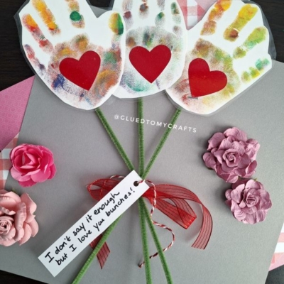 Three colorful handprint cutouts with red heart centers are arranged as flowers, with green pipe cleaner stems, pink paper flowers, and a note reading “I don’t say it enough but I love you bunches!” on a gray card.