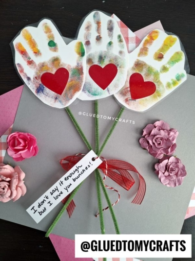 Three colorful handprint cutouts with red heart centers are arranged as flowers, with green pipe cleaner stems, pink paper flowers, and a note reading “I don’t say it enough but I love you bunches!” on a gray card.