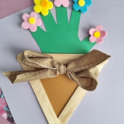 A handmade craft features a paper flower pot made from popsicle sticks, brown patterned paper, and a fabric bow, holding a green handprint cutout with colorful felt flowers attached to each fingertip.
