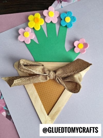 A handmade craft features a paper flower pot made from popsicle sticks, brown patterned paper, and a fabric bow, holding a green handprint cutout with colorful felt flowers attached to each fingertip.