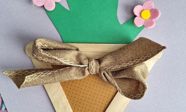 A handmade craft features a paper flower pot made from popsicle sticks, brown patterned paper, and a fabric bow, holding a green handprint cutout with colorful felt flowers attached to each fingertip.