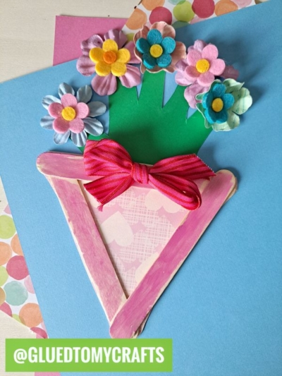 A handmade craft featuring a pink popsicle stick flowerpot with colorful paper flowers and a red bow, displayed on a blue background. @GLUEDTOMYCRAFTS is written at the bottom in a green box.