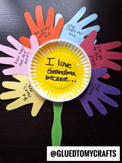 A colorful "I love mom because" flower craft made from a yellow paper plate center and handprint-shaped petals, each listing reasons for loving mom like “She cooks for me” and “She plays with me.”.