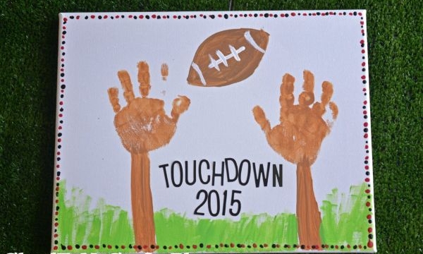 A canvas featuring two brown handprints as football goalposts, a painted football above, green grass below, TOUCHDOWN 2015 in the center, and a dotted border—perfect for HANDPRINT & FOOTPRINT KEEPSAKES.