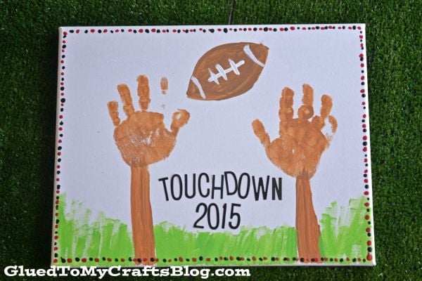 A childrens painting with two brown handprints as football uprights, a football above, TOUCHDOWN 2015 in black letters below, green grass at the bottom, and a dotted border on white paper.