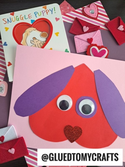 A handmade Valentine’s dog craft made from red and purple paper with large googly eyes is shown on a pink background, surrounded by craft foam heart crafts, mini felt envelopes, and the book Snuggle Puppy! @GLUEDTOMYCRAFTS is at the bottom.