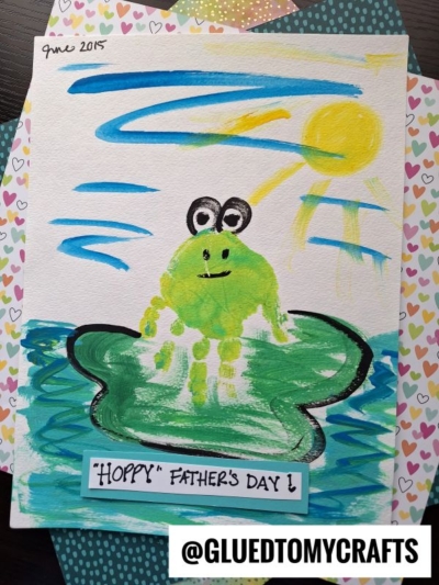 A HANDPRINT FATHER'S DAY painting of a green frog on a lily pad in blue water under a yellow sun. Text says “HOPPY FATHERS DAY!” and “@GLUEDTOMYCRAFTS.” Heart-patterned papers decorate the background.
