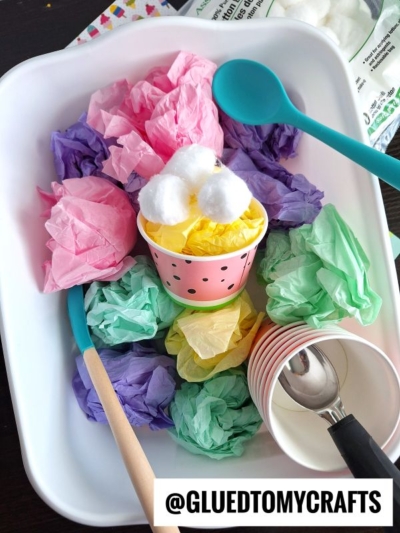 A sensory bin with crumpled tissue paper in pastel colors, a pink paper cup filled with yellow tissue and cotton balls, plastic spoons, and stacked cups. The handle @GLUEDTOMYCRAFTS is labeled at the bottom.