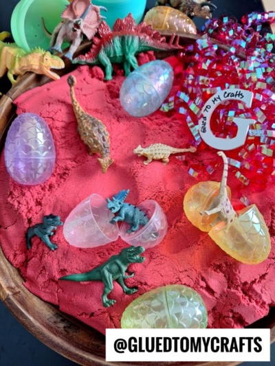 A sensory play tray with red sand, plastic dinosaur toys, iridescent egg-shaped containers, and colorful shredded paper. A label reads @GLUEDTOMYCRAFTS.