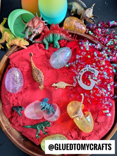 A sensory play tray with red sand, toy dinosaurs, plastic dinosaur eggs, shredded red foil, and a circular tag labeled Glued To My Crafts on a wooden tray.