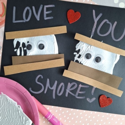 A spatula stamped smores craft card with LOVE YOU SMORE! written in chalk on black paper, featuring two smores made from brown strips, white paint, googly eyes, and red glitter hearts. Craft supplies are scattered around the card.